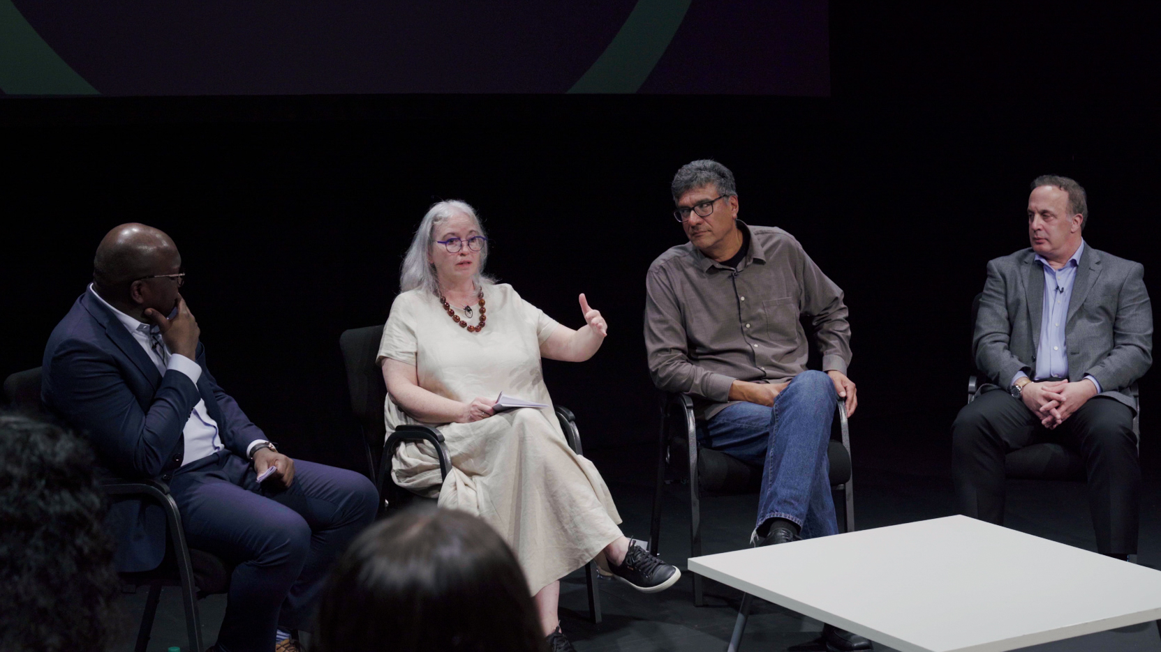 Four McMaster experts sit in chairs, addressing an audience out of frame. A black theatre backdrop is behind them. Panelists listen to panel member second from the left, dressed in white, as she speaks and gestures to audience.