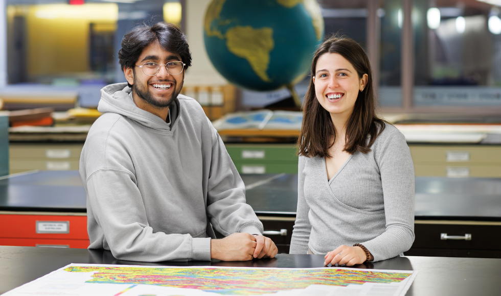 Two people stand behind a large table that has a map on it while smiling at the camera. Behind them is a large globe and an open book containing maps.