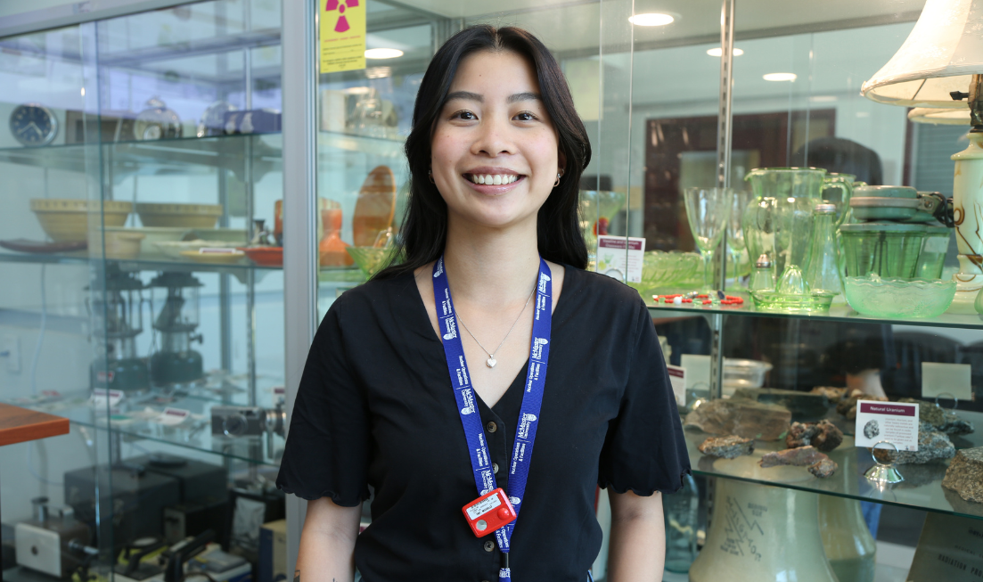 A person with long dark hair in a dark T shirt and a lanyard smiling in front of a wide glass-fronted case full of small items: rocks, glassware, bowls.