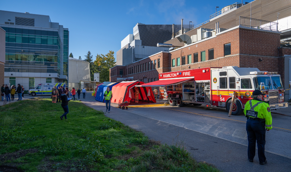 An ambulance, firetruck, and blue and red tents on McMaster's campus. There are people, some in fluorescent safety vests, walking around.