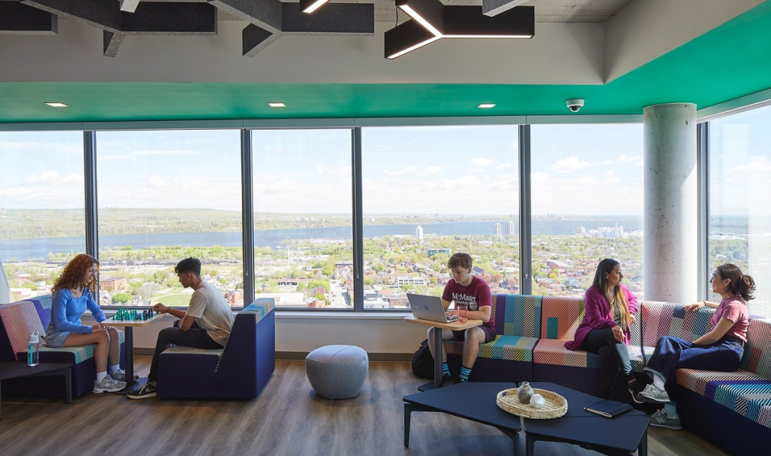 Alt Text: Against a backdrop of Hamilton’s skyline and Lake Ontario, students sit on couches playing chess, chatting or working on their laptop in the Sky Lounge high up in the 10 Bay residence building.