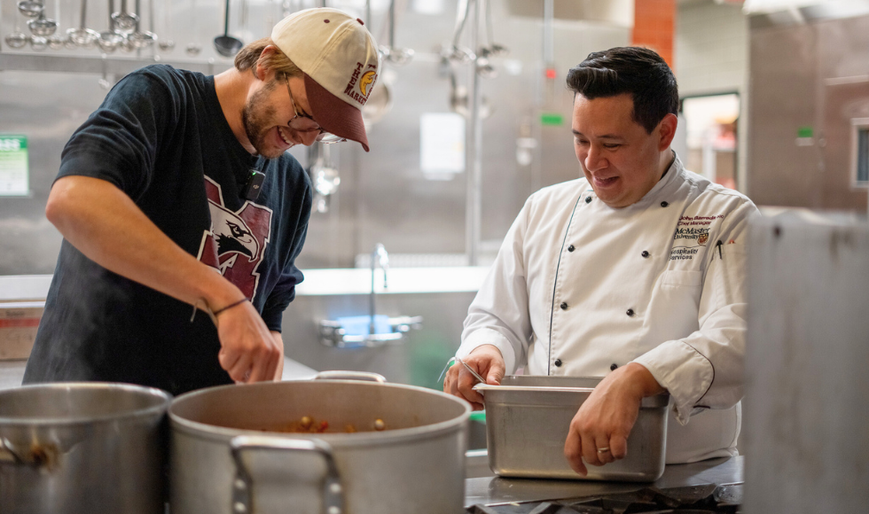 A student in a Marauders t-short and a ball cap leaned over a stove cooking while another person wearing chef’s whites looks on