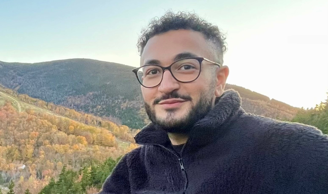 Head and shoulders of a young person with glasses and a beard and moustache in a black sweatshirt in the foreground. In the background are hills and a blue sky.