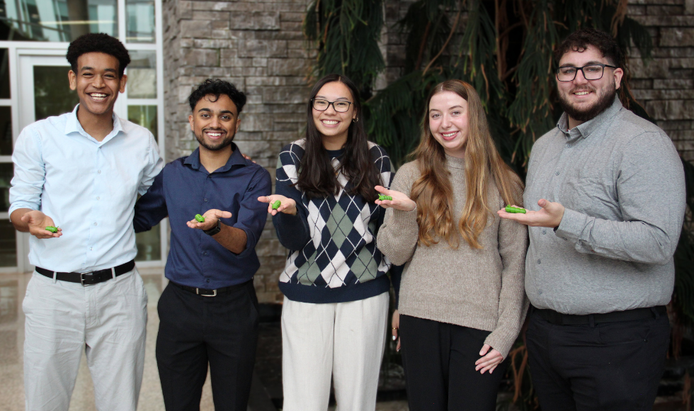 Five smiling MBDC students stand side-by-side, each holding a small green object, the 