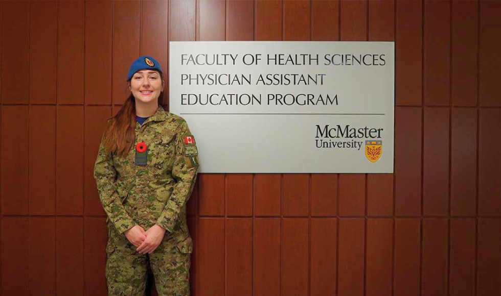 Officer Cadet Marissa Nociar wearing a Canadian Forces military field uniform while standing beside a sign that reads, ‘Faculty of Health Sciences Physician Assistant Education Program.’ The sign bears the logo of McMaster University.