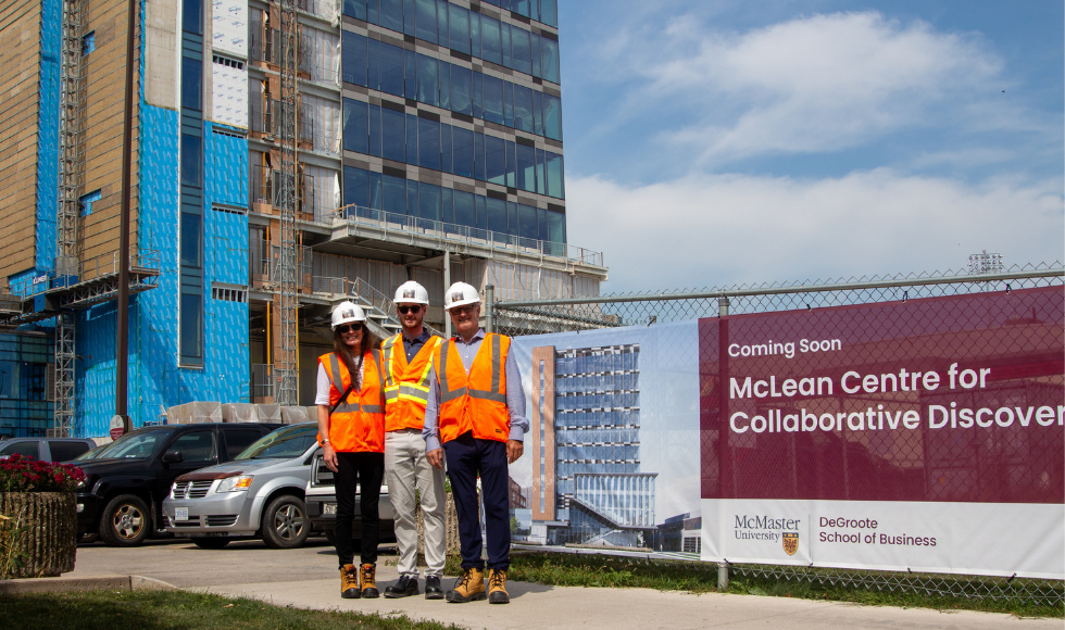 Three people in hard hats, reflective vests and steel-toed boots at a construction site, with a sign that says 