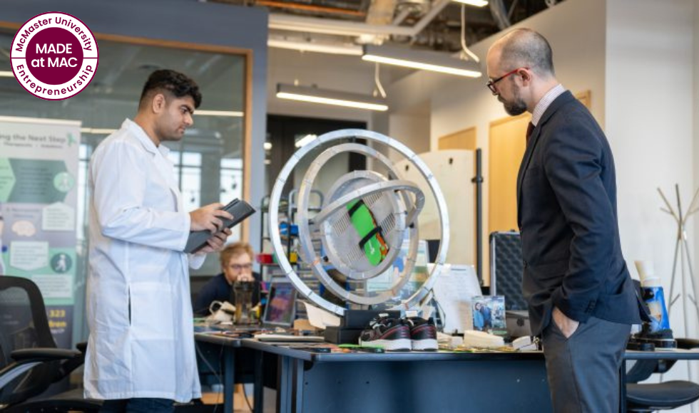 A bearded person in a suit and tie and a person in a lab coat look at a large piece of science equipment on a table in a lab with . A graphic over top reads: Made at Mac: McMaster Entrepreneurship.