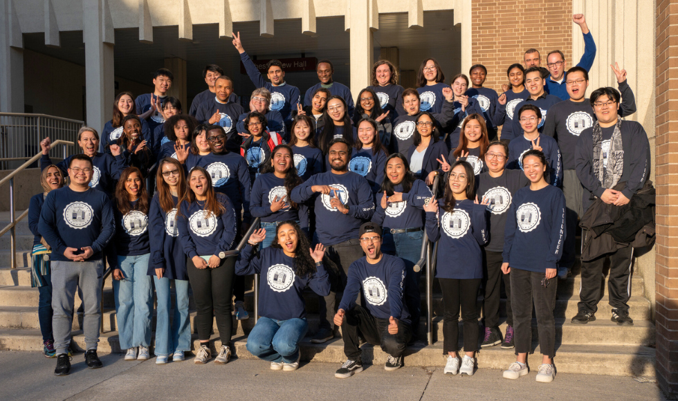 A large group of students standing on an outdoor staircase. They are all wearing the same navy blue, long sleeve shirt that has text on it that reads, ‘Tax Squad - Helping Hamilton.’