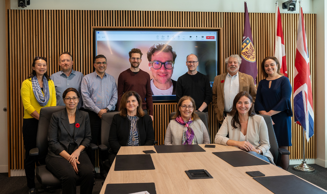 A group of smiling people arranged in two rows— front row is four people sitting at the end of a table, back row has seven people standing, with space between them for a large monitor on the wall, showing the face of a person who joined the group by a video call.