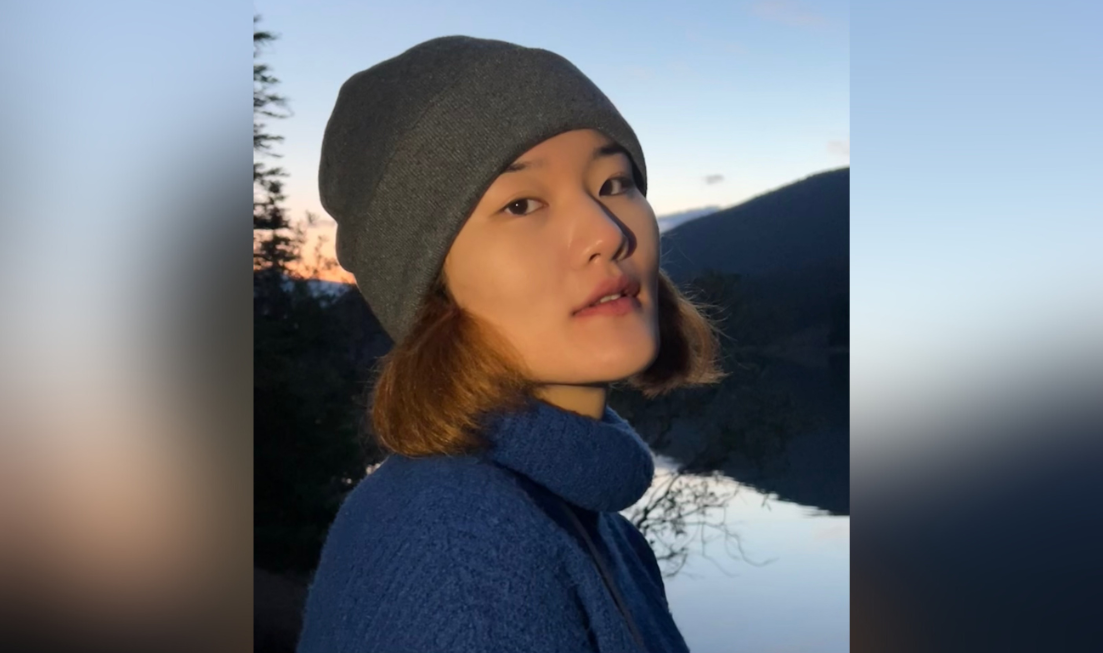 Headshot of a person turned sideways but looking at the camera, wearing a toque and a turtleneck sweater, with a lake, mountain and twilight in the background.