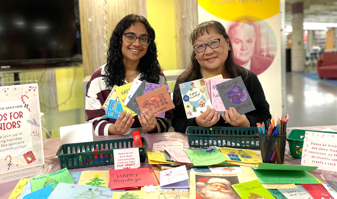 Poornima and Ann, both with long black hair and glasses, sit at a table in H.G. Thode Library, surrounded by art supplies and cards, and holding cards made by students.