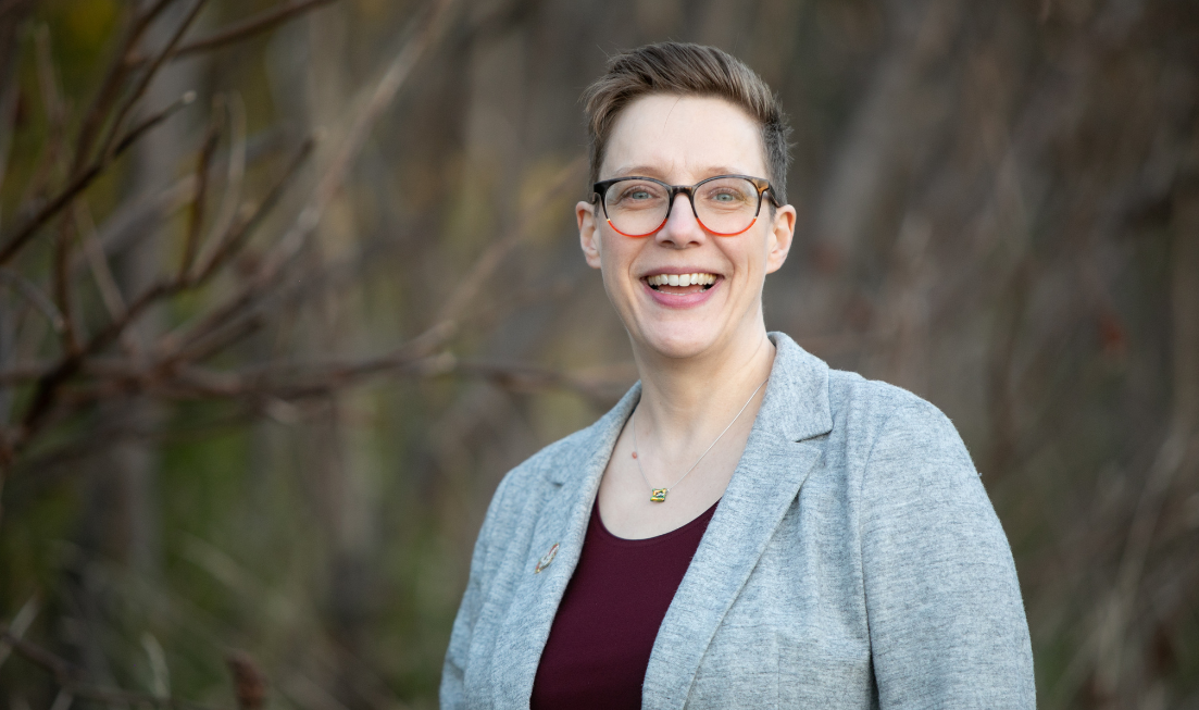 Head and shoulders of smiling Catherine Anderson standing outside. she has light skin, short, dark hair and dark-framed glasses. She is wearing a maroon top with a light grey blazer over it.