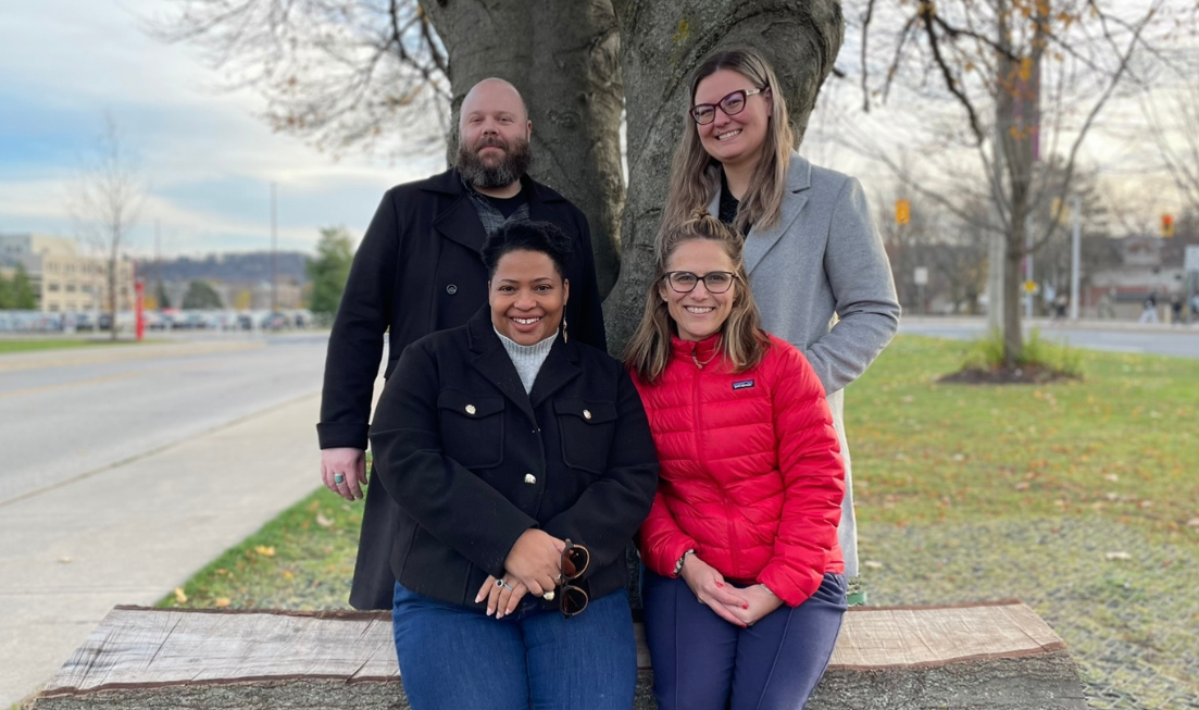 Crystal with short black hair and a black coat and Lynne with long blonde hair and a red coat sit on a wooden bench outside. Casey with a shaved head and a black coat and Anna with long blonde hair and a grey coat stand behind Crystal and Lynne.