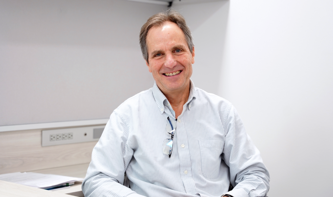 Upper-body image of smiling Jerry Hurley sitting at a desk. He has short, dark hair and is wearing a light-coloured button-up shirt with a pair of glasses hanging from the neckline.