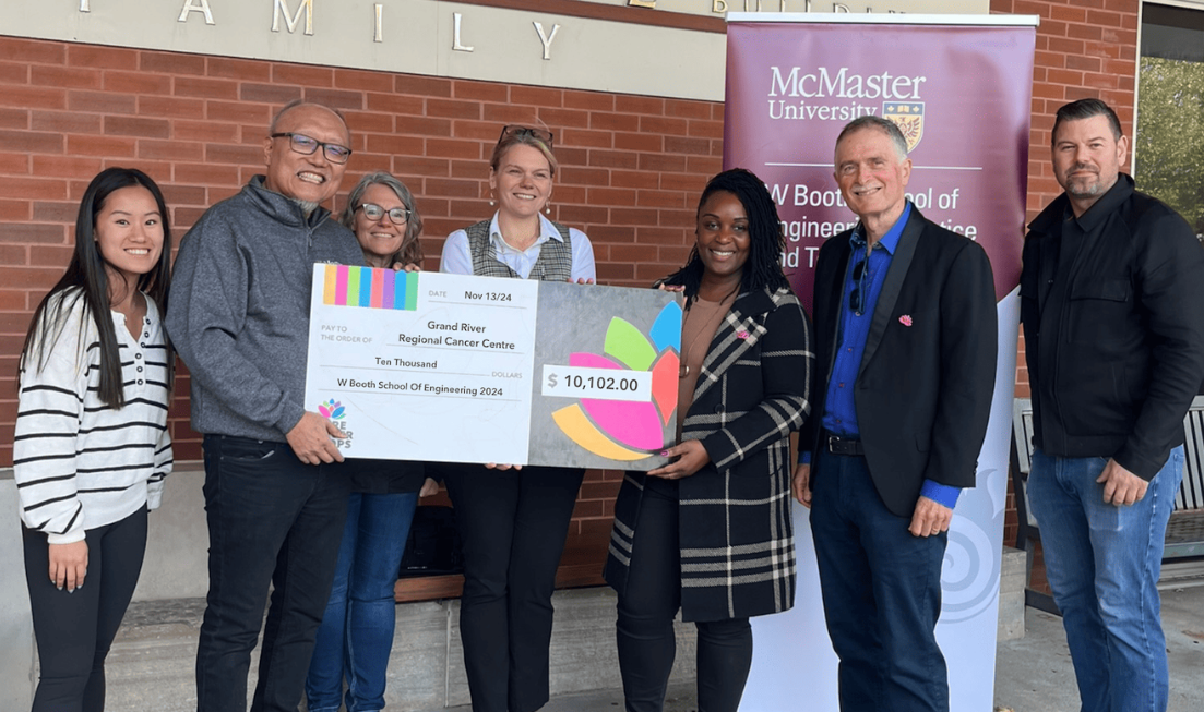 Six people stand in a row, outside hospital building, smiling. Two are holding up a giant cheque for $10,102, and in the background is a banner that reads W Booth School of Engineering Technology and Practice.