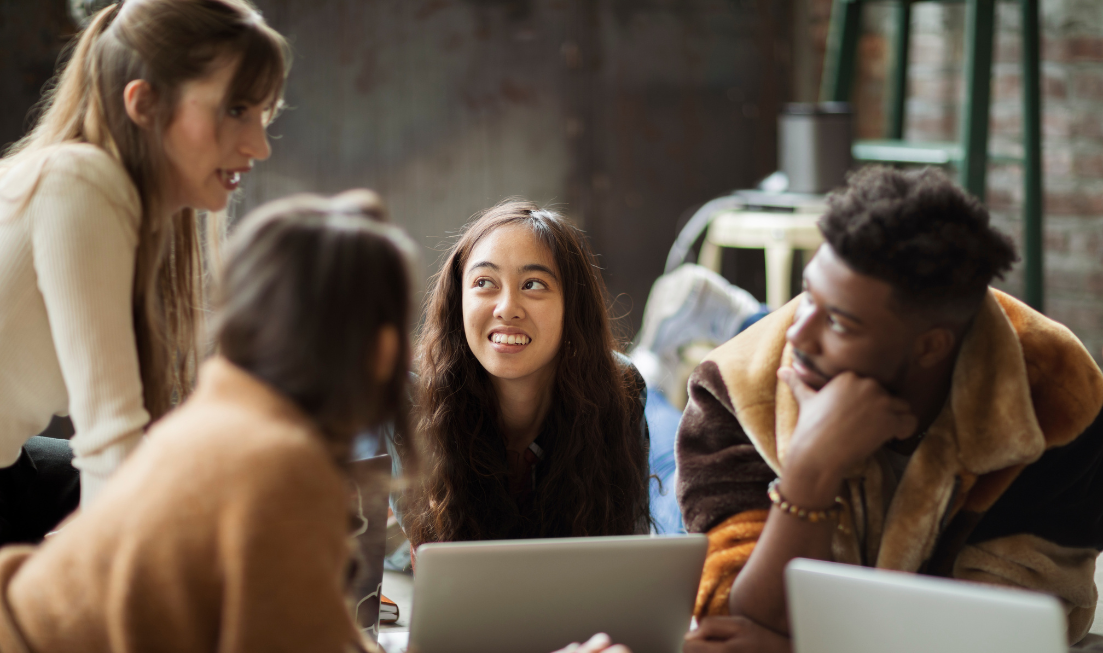 Four cheerful-looking young people sitting and standing around a desk with a laptop on it.