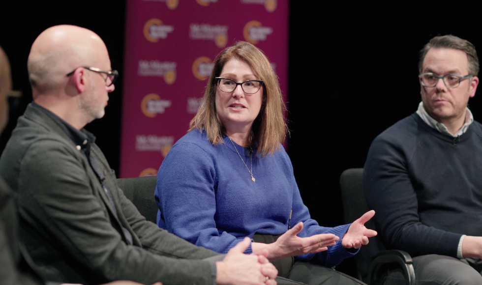 Three people seated. The person in the centre is speaking while the other two look on. There is a banner behind them that has McMaster University branding on it.