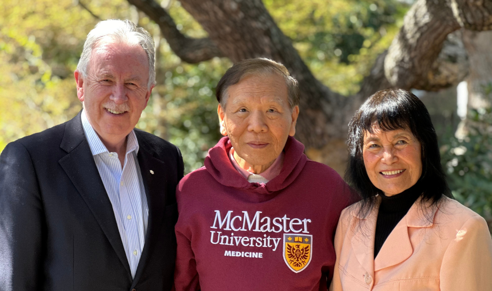 Three people stand for a photo. The person in the middle is wearing a maroon sweater with the McMaster University Medicine logo.