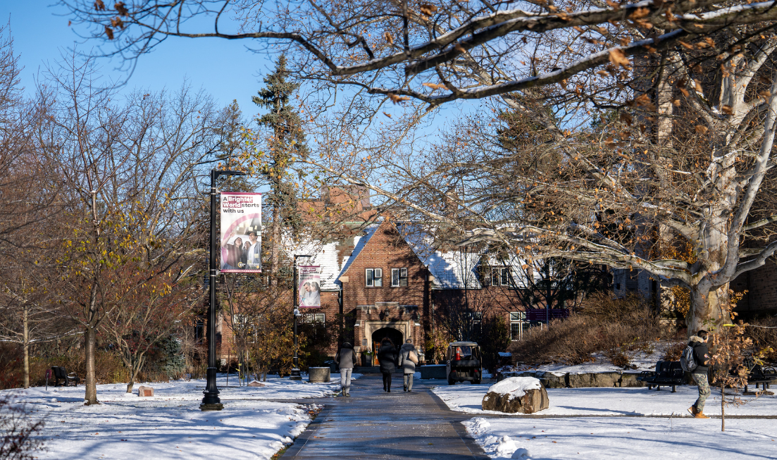 Seen from behind, a few people walking down a cleared path on campus on a sunny, snowy day, with a campus building in the background.