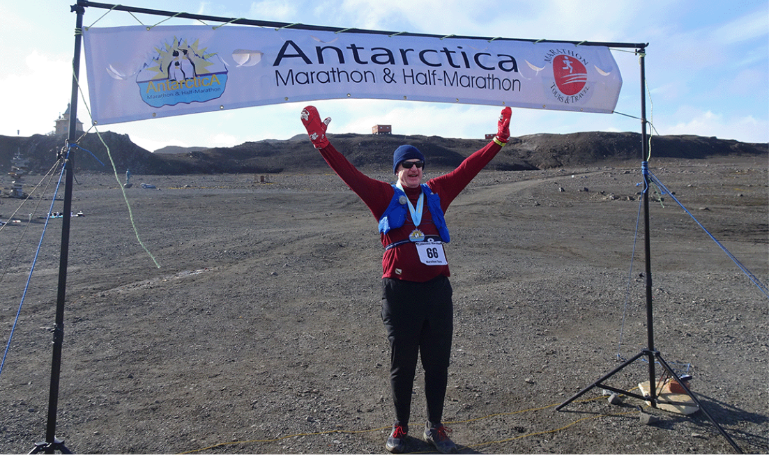 A smiling Mark Walton wearing running gear, with a medal around his neck, stands with his arms outstretched under the banner for the Antarctica marathon and. half-marathon.