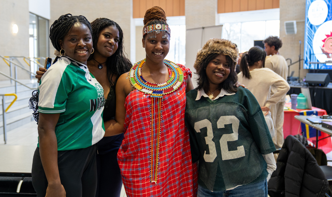 A student in traditional Maasai clothes and jewelry smiles with three friends in the atrium of MUSC.