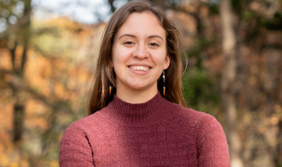 A young woman, standing outdoors with trees behind her, smiles at the camera.