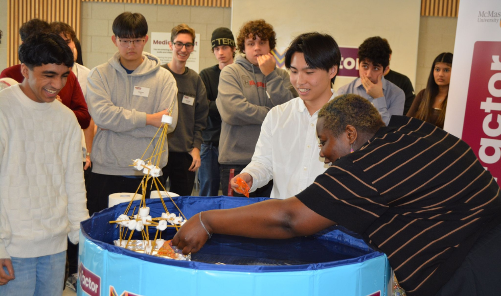 Several university students stand around a large blue tub, within which they are building a structure out of marshmallows and uncooked spaghetti. A few students are leaning into the bin, building and laughing, while other students stand and watch.