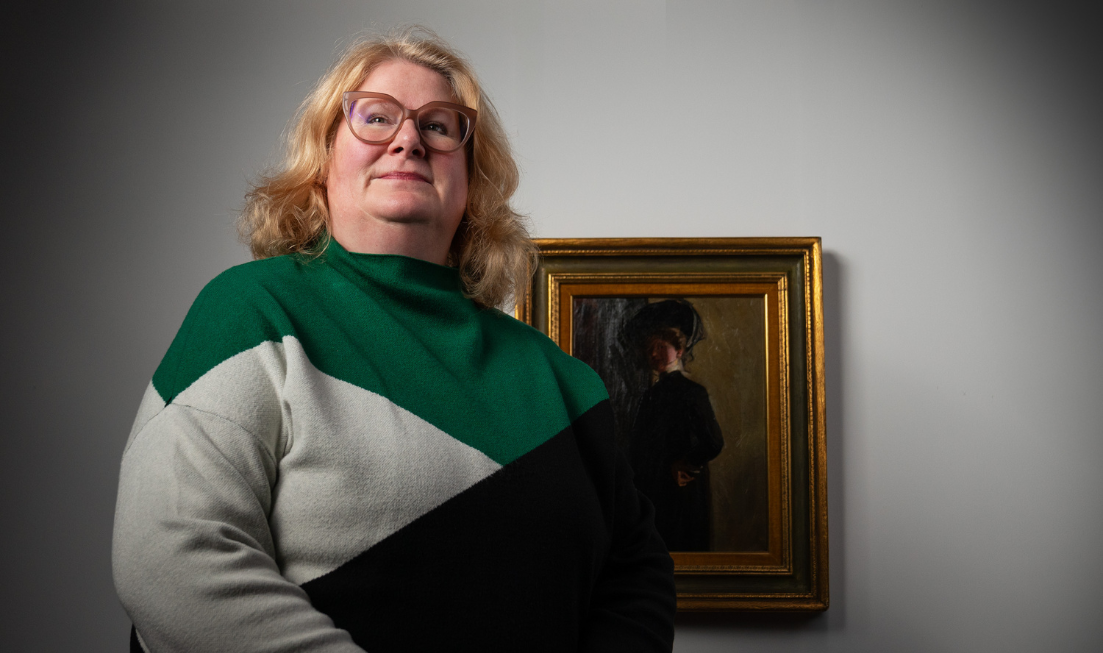 Waist up image of Mary Reid, with blonde hair, red-framed glasses and a colour-block sweater, standing obliquely in front of a framed painting on a light-coloured wall.