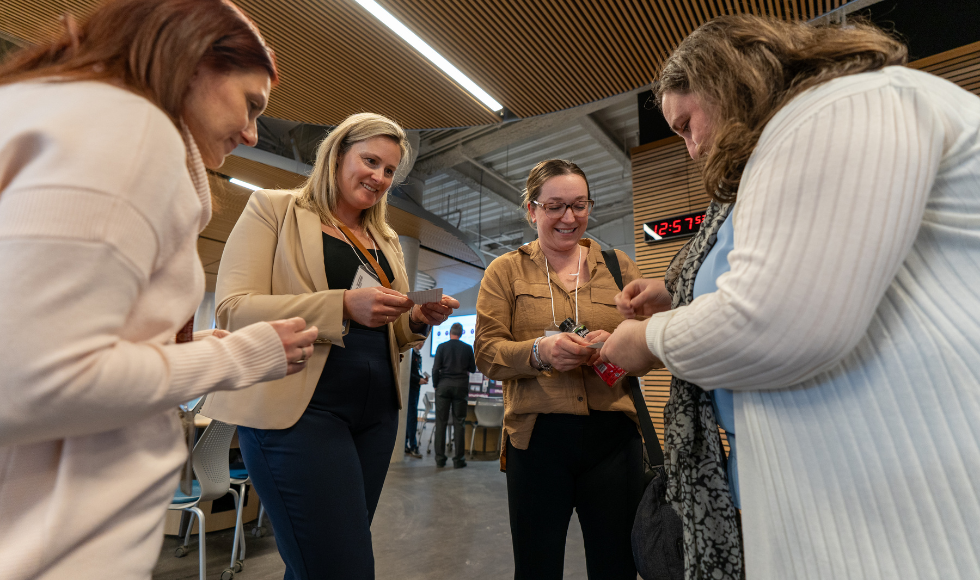 Inside of a large hall, four women stand in a loose circle, facing each other. Two of them are holding small pieces of paper out towards a third woman, who appears to be writing something down.