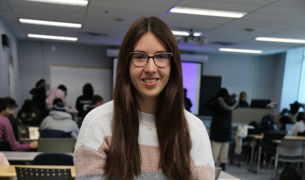 A woman wearing glasses smiles, facing the camera. Her head and shoulders are visible. Behind her is a classroom, with several more people further away, seated and facing away.