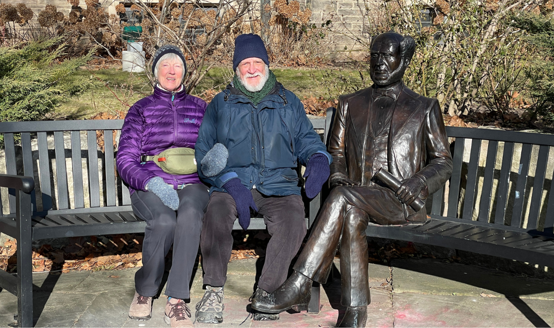 A white-haired woman and a white-haired man, both smiling and wearing winter clothes, sit beside the statue of Senator McMaster on campus.