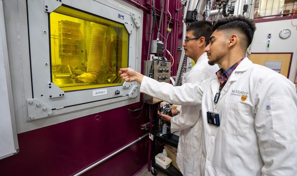 Two young men in lab coats stand beside a large red structure that has a yellow window. One person has a hand raised, gesturing at the yellow window.