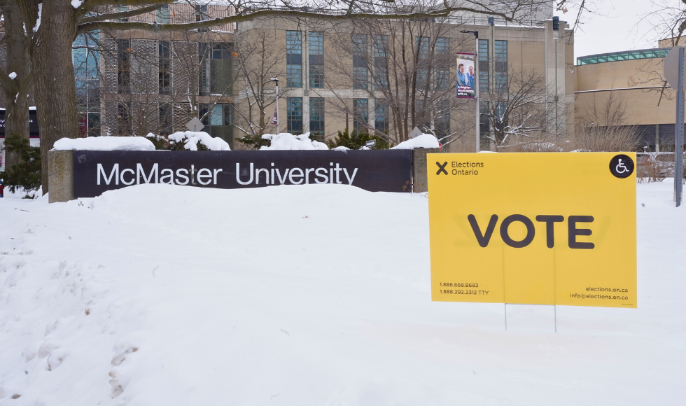 A yellow lawn sign that reads 'VOTE' sits on a snowy lawn in front of a stone building, with a McMaster University sign just in front of the building.