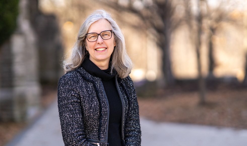 Smiling and shoulders of Andrea Farquhar in a dark blazer and top, standing outdoors on campus.