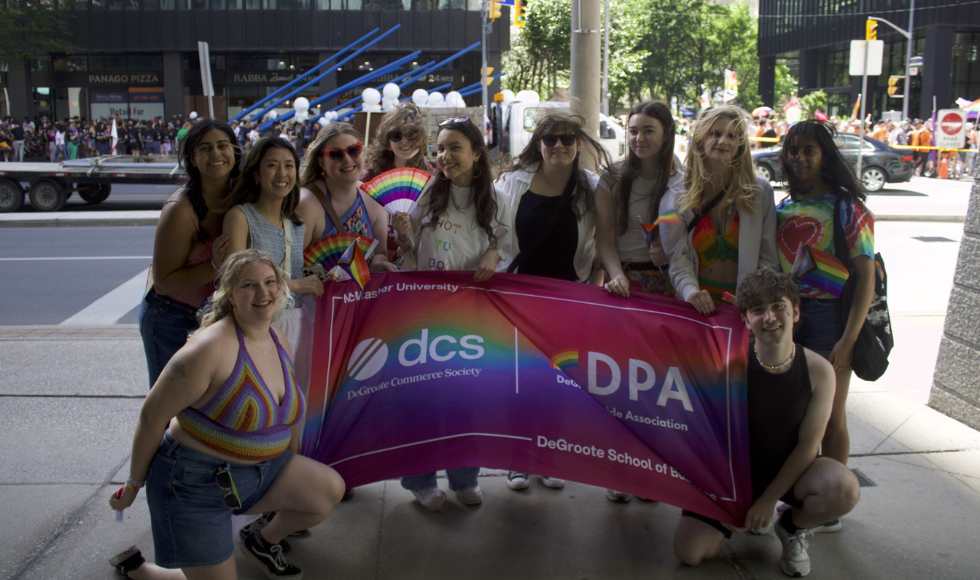 A group of people, several dressed in rainbow shirts or holding rainbow fans, stand on a city sidewalk. Tthey are holding up a large banner that reads 'McMaster University - DeGroote Commerce Society - DeGroote Pride Association - DeGroote School of Business.