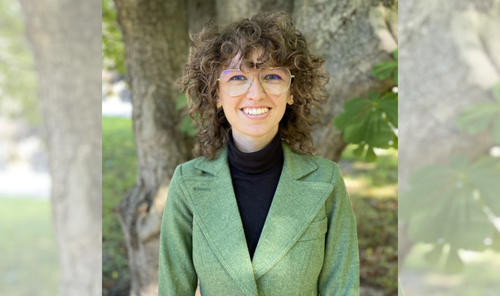 A headshot of a woman, wearing glasses and a green blazer. She is standing outside, with a tree behind her.