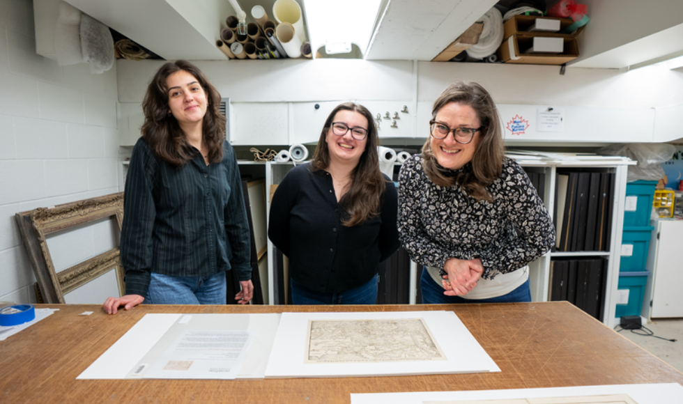 Three women stand in front of a large wooden table, which has a large map laid out on it. They are all smiling at the camera.