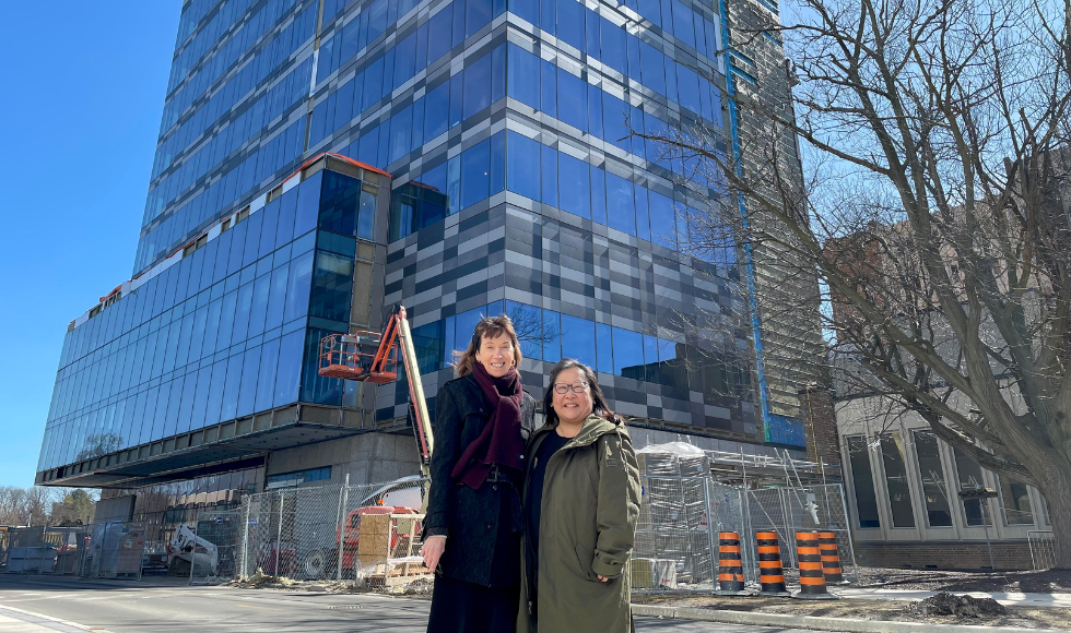 Two people, wearing winter coats, stand in front of a new building under construction. A crane and construction fencing, near the base of the building, is visible behind them.