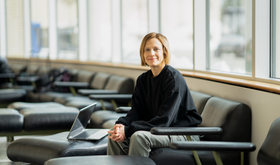 A woman seated on a leather couch with her elbows on her knees and her hands clasped together. She is looking directly at the camera and there is an open laptop beside her.