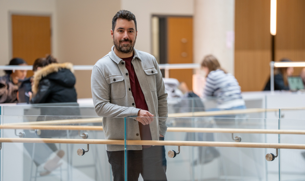 Ryan Rexworthy stands in a building, along a clear walkway, resting his arm on the railing. Several students are visible sitting in the background.