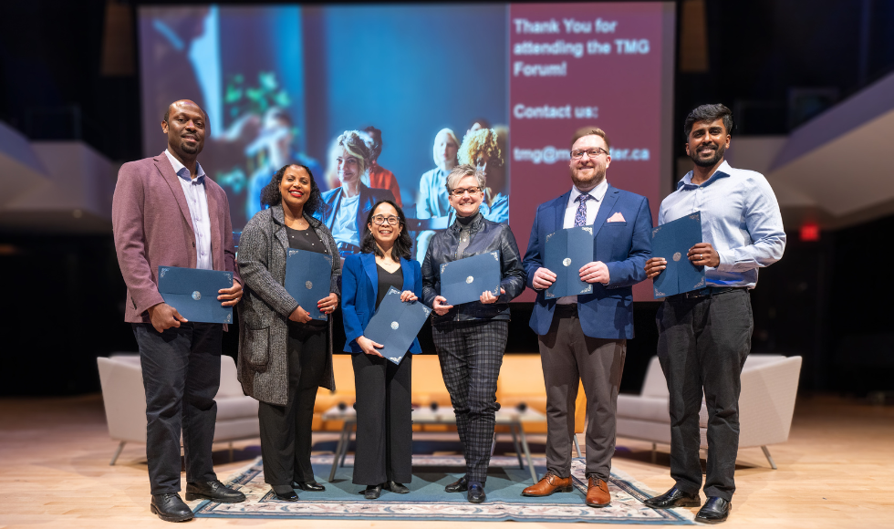 Six people standing on a stage. They are all holding large blue envelopes and smiling at the camera.