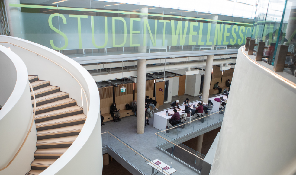 The interior of a building atrium, with large glass windows at the front. A curved staircase is on the left, and students can be seen below.