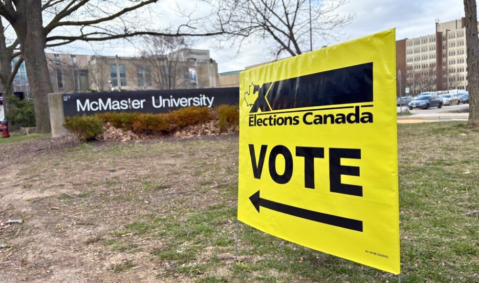 An Elections Canada polling station sign in the foreground pointing toward the McMaster University sign at the entry to campus.
