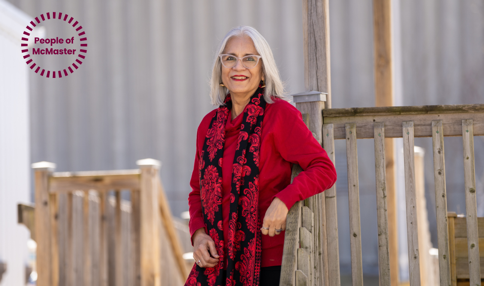 Rukhsana Jamal, who has shoulder-length grey hair and glasses, wearing red and black and leaning against a wooden rail outdoors. In the corner is a 