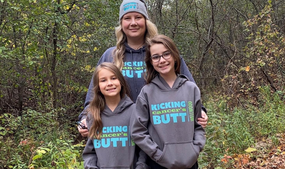 A woman and two young girls stand in front of a green forest. They are all wearing matching grey sweatshirts that read 'Kicking Cancer's Butt.'