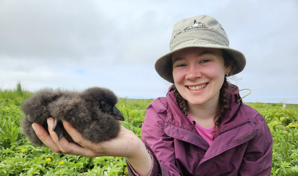 A woman in a hat holds a fluffy black bird chick in her outstretched hand.
