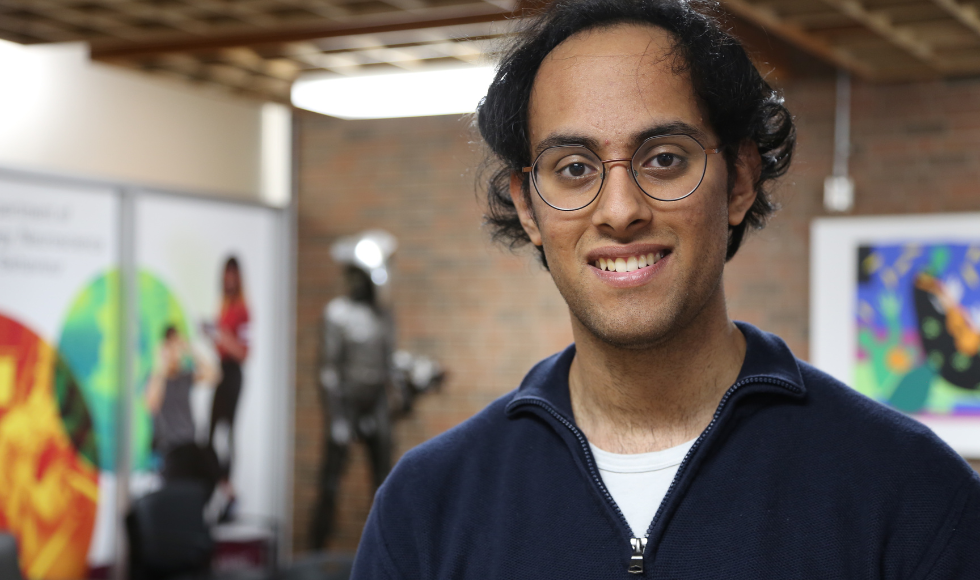 A man in a blue sweater and glasses stands in a hallway, with colourful murals and a brick wall visible behind him.