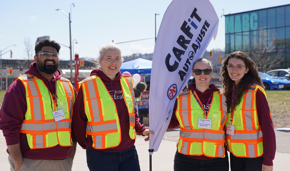 Four people in safety vests stand outside in a parking lot, posing for the camera and holding a banner that says 'Carfit.'