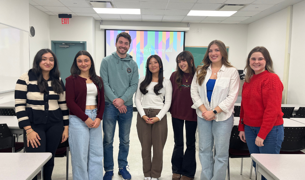 Seven people stand in a classroom, all facing the camera and smiling.