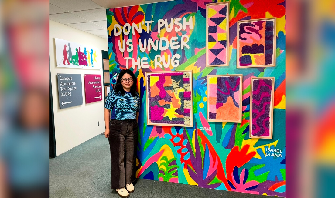 Isabel Diana stands beside her vibrant and colourful artwork 'Don't Push Us Under The Rug' at Mills Memorial Library.